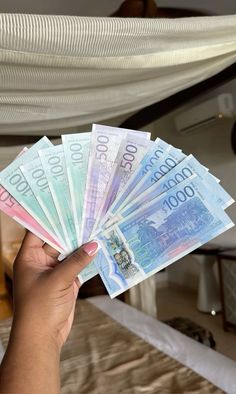 a person holding five bills in their hand on top of a bed with white sheets