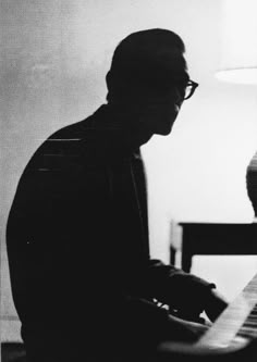 a black and white photo of a man playing the piano in front of a lamp