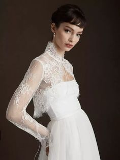 a woman wearing a white dress with sheer sleeves and lace on the top, standing in front of a dark background