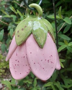 a pink and green flower shaped ornament hanging from a tree branch in front of some bushes