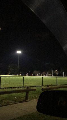 a baseball field at night with lights on and people playing in the grass behind it