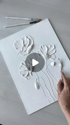 someone is making an art project with white paper and flowers on the board, while another person holds a paintbrush