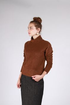 a woman standing in front of a white wall wearing a brown sweater and black skirt