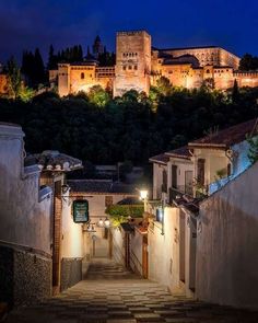 an alley way leading to a castle at night with lights on the buildings and trees in the background