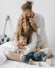 a man and woman are sitting on a bed with a baby in their lap while the child is being held up to his ear