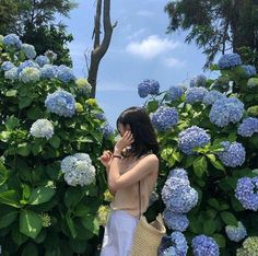 a woman standing in front of blue flowers talking on her cell phone while holding a straw bag