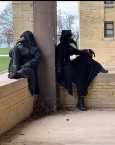 two people dressed in black sitting on the side of a building with their backs to each other