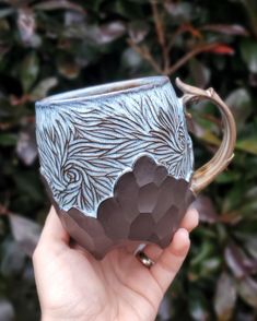 a hand holding a blue and white coffee mug in front of some green bushes with leaves on it