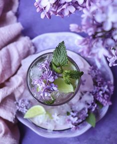 a glass filled with ice and lime next to purple lilacs on a plate,