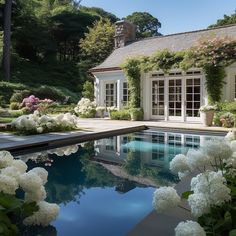 a pool surrounded by white flowers next to a house with large windows and lots of greenery