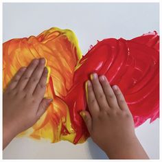 two hands are painting an orange and yellow piece of art with red paint on white paper