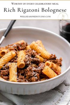 a white bowl filled with pasta and meat on top of a table next to a glass of wine