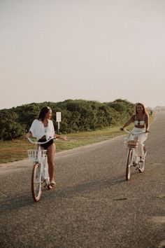 two women riding bikes down the middle of an empty road with trees in the background