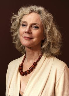 a woman with blonde hair wearing a white cardigan and beaded necklace is looking at the camera