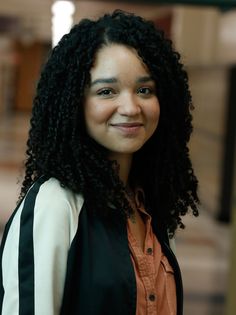 a woman with long curly hair smiling at the camera while wearing an orange shirt and black vest