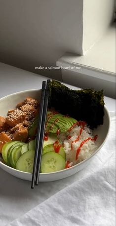 a white bowl filled with rice, cucumber and meat next to chopsticks