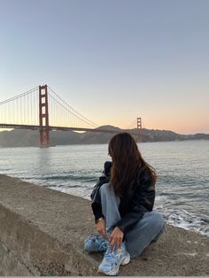 a woman sitting on the edge of a concrete wall next to water and a bridge