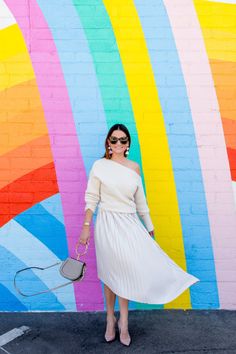 a woman standing in front of a colorful wall wearing a white dress and holding a gray purse