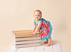 a baby girl in a red and blue dress standing next to a wooden crate on a white floor