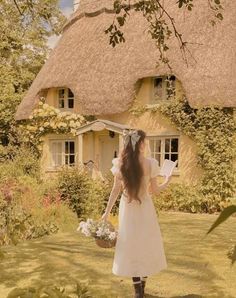 a woman in a white dress holding a basket and walking towards a thatched house