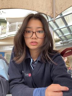 a woman wearing glasses sitting at a table with an umbrella over her head and looking off to the side