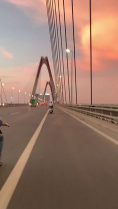 a man riding a skateboard down a street next to a tall metal bridge at sunset