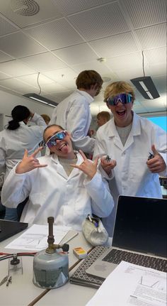 two men in lab coats and goggles standing next to each other near a laptop