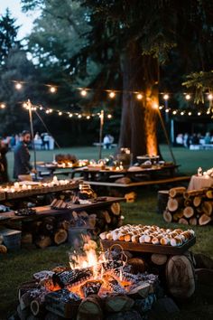 an outdoor fire pit with food on it and string lights strung over the trees in the background