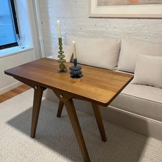 a wooden table with two candles on it in front of a white couch and window