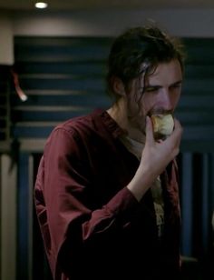 a man with long hair eating a donut in a dark room next to a radiator