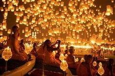 many people are sitting on the ground and looking at paper lanterns in the night sky