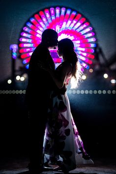 a couple standing next to each other in front of a colorful ferris wheel at night