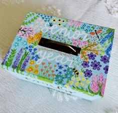 a tissue box with flowers and butterflies painted on the front, sitting on a bed