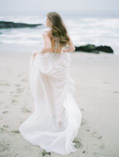 a woman in a white dress is walking on the sand at the beach with her back to the camera