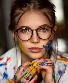 a woman with glasses and paint on her face is looking at the camera while she has her hands painted in different colors