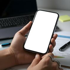 a person holding a cell phone in their hand while sitting at a desk next to a laptop