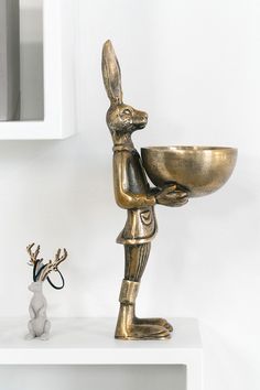 a statue of a rabbit holding a bowl on top of a white shelf next to a small figurine