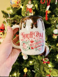 a hand holding a coffee mug with the north pole hot chocolate on it in front of a christmas tree