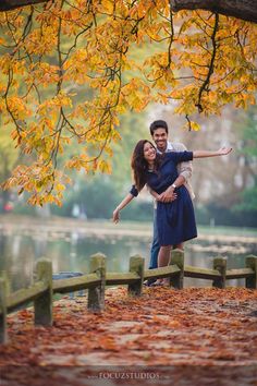 a man and woman standing on a bridge in front of a tree with yellow leaves