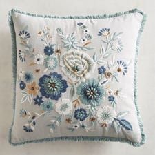 a white pillow with blue flowers and fringe trimmings on the front, sitting against a wall