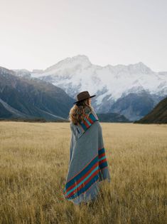 a woman wearing a blanket and hat standing in a field with mountains in the background