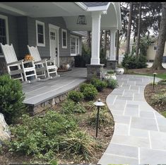 a porch with two white rocking chairs on it and landscaping in the front yard area