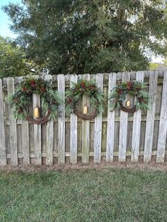 three christmas wreaths hanging on a wooden fence with lights in the middle and greenery around them