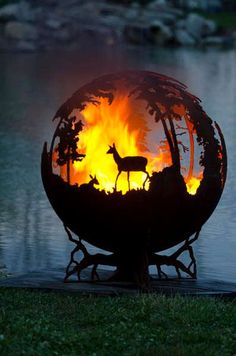 an outdoor fire pit with deer and trees in the center, on top of grass next to water