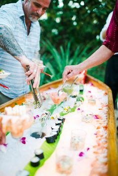 two men are serving sushi to each other on a long wooden table with white cloth