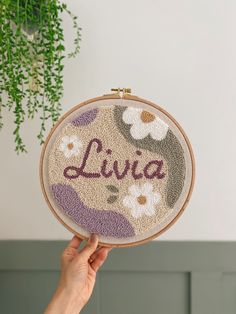 a hand holding up a small embroidered hoop with the word livva on it in front of a plant