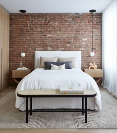 a bed with white sheets and pillows in front of a brick wall, next to a coffee table