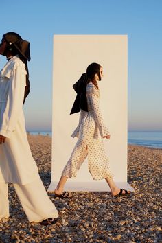 two women walking on the beach with one wearing a black hat and white jumpsuit