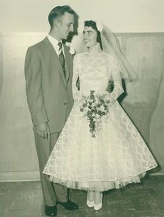 an old black and white photo of a man and woman dressed in wedding attire standing next to each other