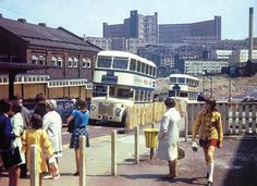 several people are standing on the sidewalk near two double decker buses and some buildings in the background